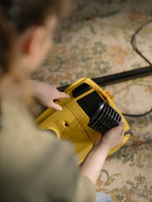 A person holding a yellow portable carpet cleaning or dry vacuum machine in a residential setting, standing on an intricately patterned rug with shades of beige, green, and orange. The machine feature