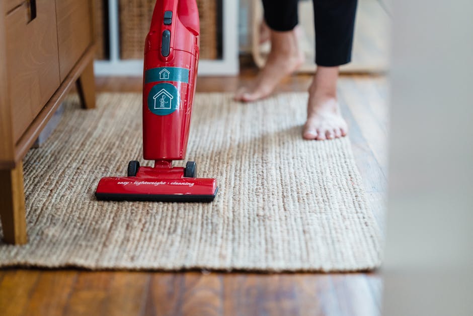 A woman with curly hair, wearing a beige jacket, blue jeans, and white shoes, is performing surface cleaning with a vacuum cleaner on a patterned beige and blue rug in a residential living room. The r