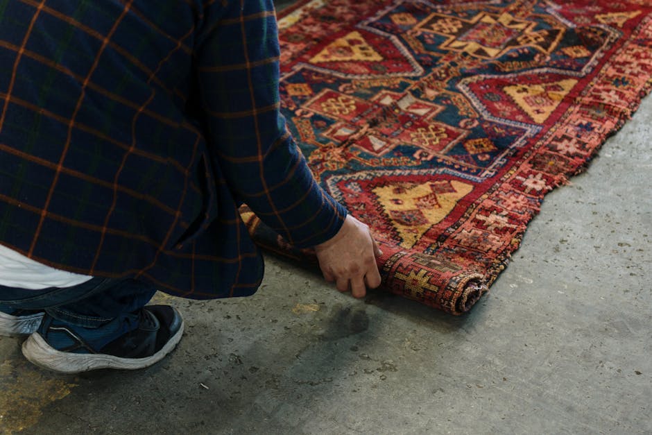 Close-up image of a decorative area rug resting on a wooden floor. The rug features a detailed floral and geometric pattern with warm tones of red, cream, and blue, and has fringed edges that appear s